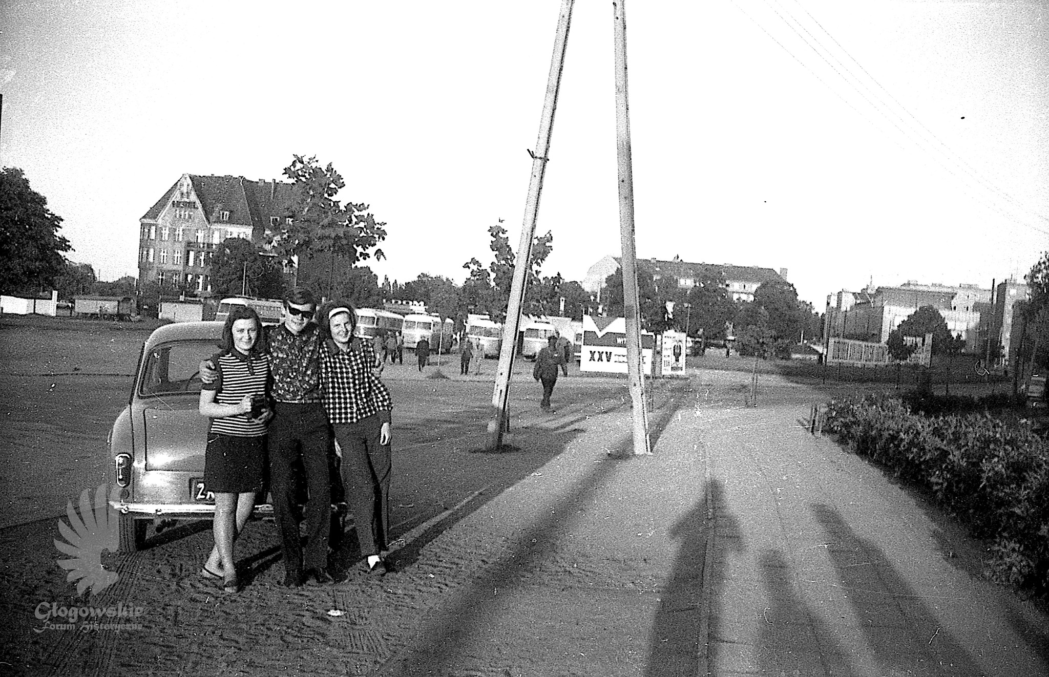 Młodzi ludzie pozujący do zdjęcia przy samochodzie marki Syrena. W tle stary plac dworcowy z autbusami marki Robur